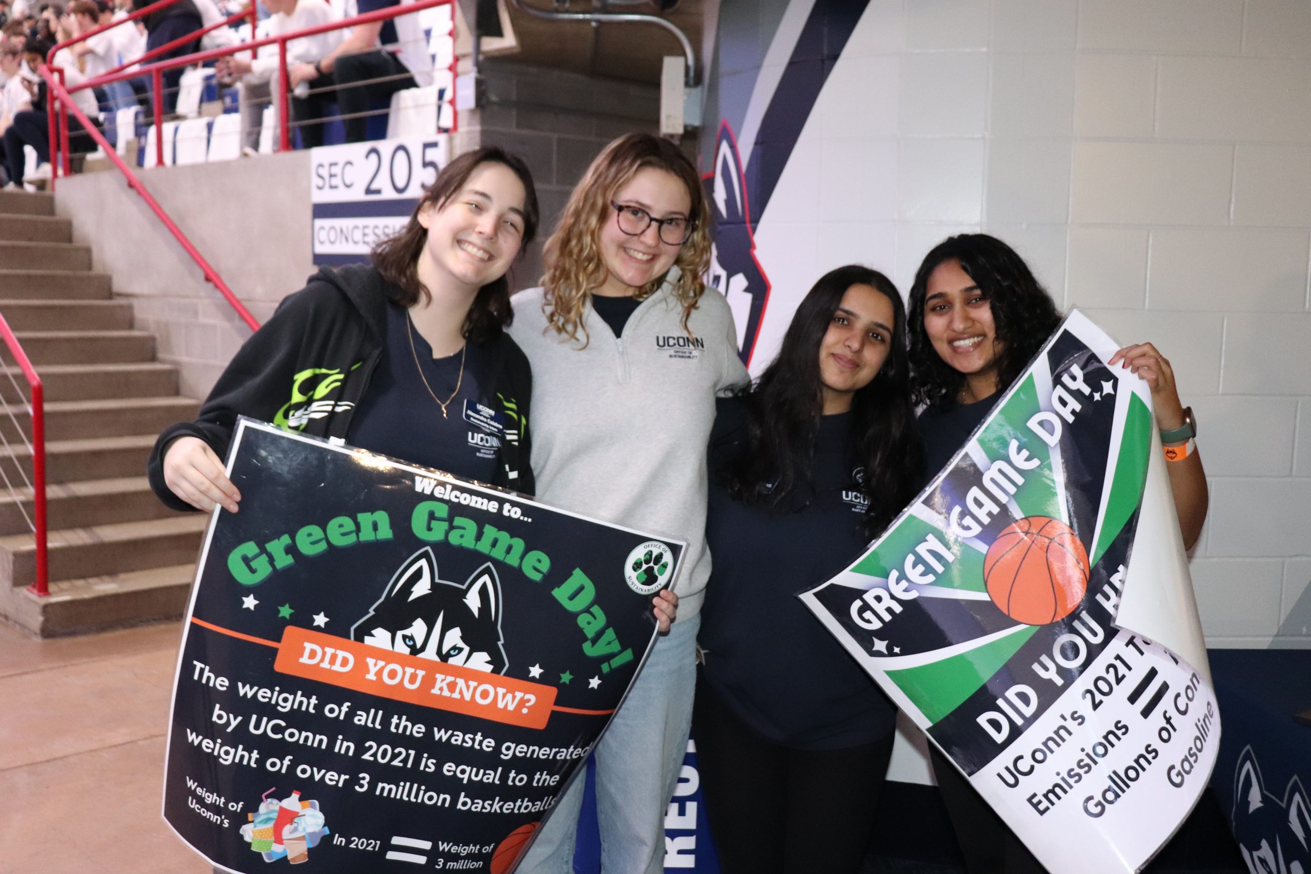 Four OS interns smiling and holding "Green Game Day" signs at Gampel Pavilion. The visible sign reads, "Welcome to Green Game Day. DID YOU KNOW? The weight of all the waste generated by UConn in 2021 is equal to the weight of over 3 million basketballs."