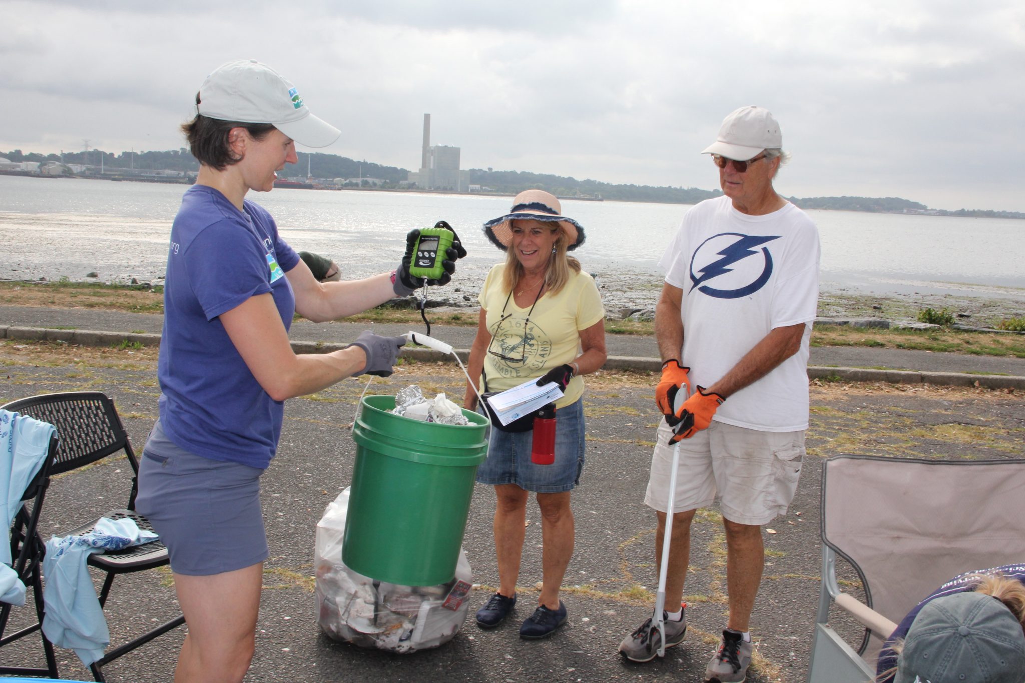 Three people are weighing a trash-filled bucket during a waterfront cleanup