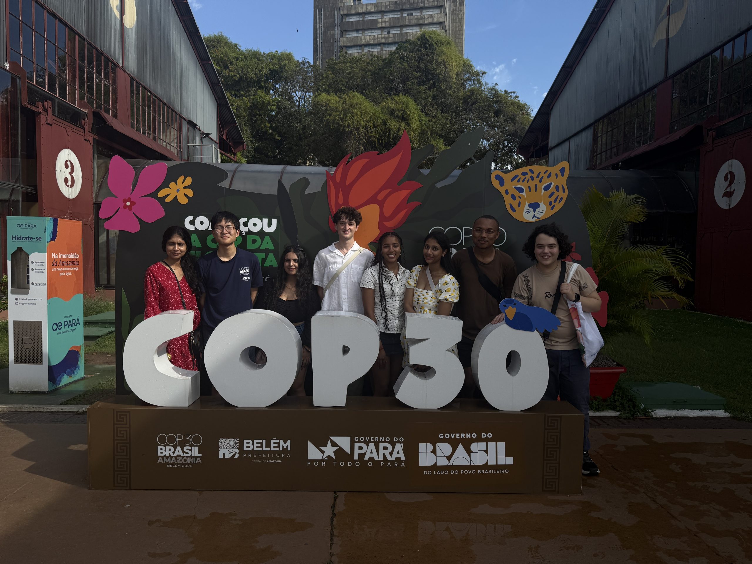 Group of UConn students (UConn@COP fellows) standing around a "COP 30" sign, with floral decorations in the background"